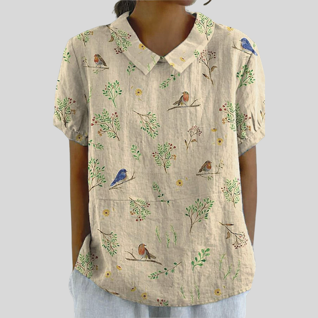 A woman wearing a graceful cream top printed with little birds perched among leafy branches, with a refined collar at an afternoon celebration.