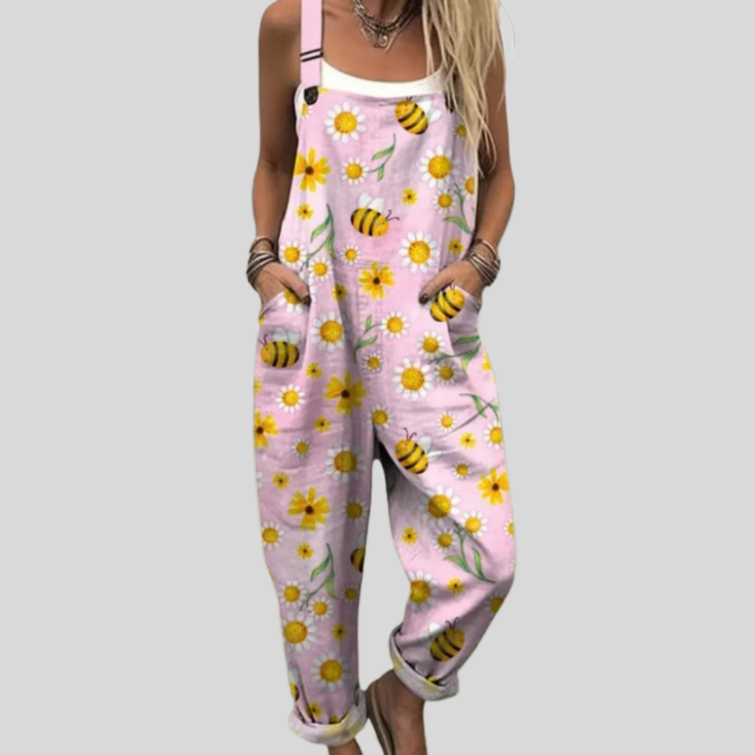 Woman wearing soft pink overalls printed with bees and yellow daisies, standing relaxed and smiling.