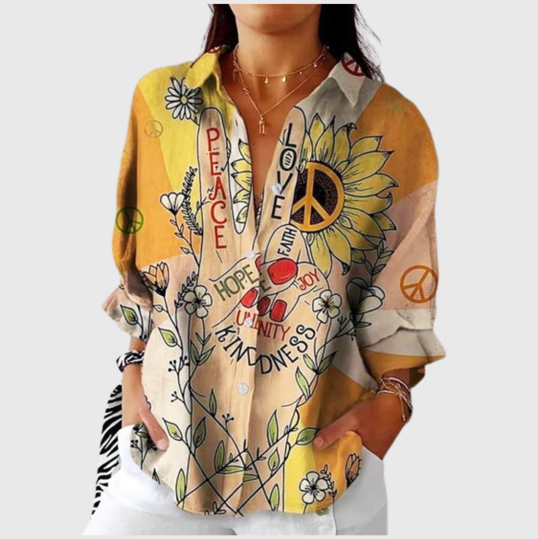 A woman wearing a warm-toned blouse with a peace-sign and sunflower graphic print and uplifting words, worn open over white pants.