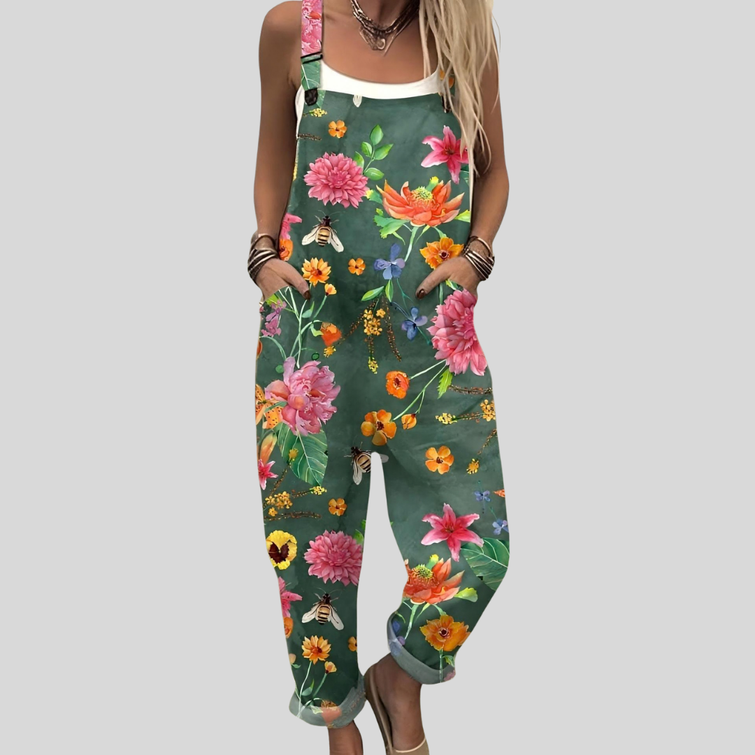 A woman wearing deep teal green overalls with a lush pink, orange and yellow floral print, smiling on a warm day.