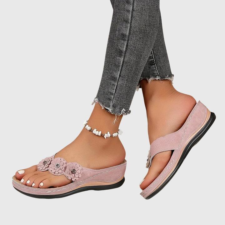 A woman wearing tan flower-trim flip-flops with a toe-post front and a low wedge sole.