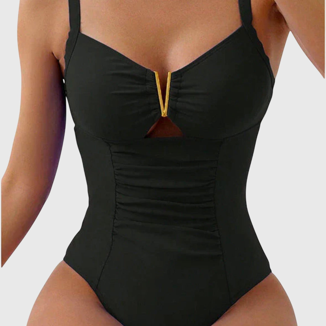 A woman by the water in a plain blue V-neck swimsuit with a small gold detail, poised in the summer light.