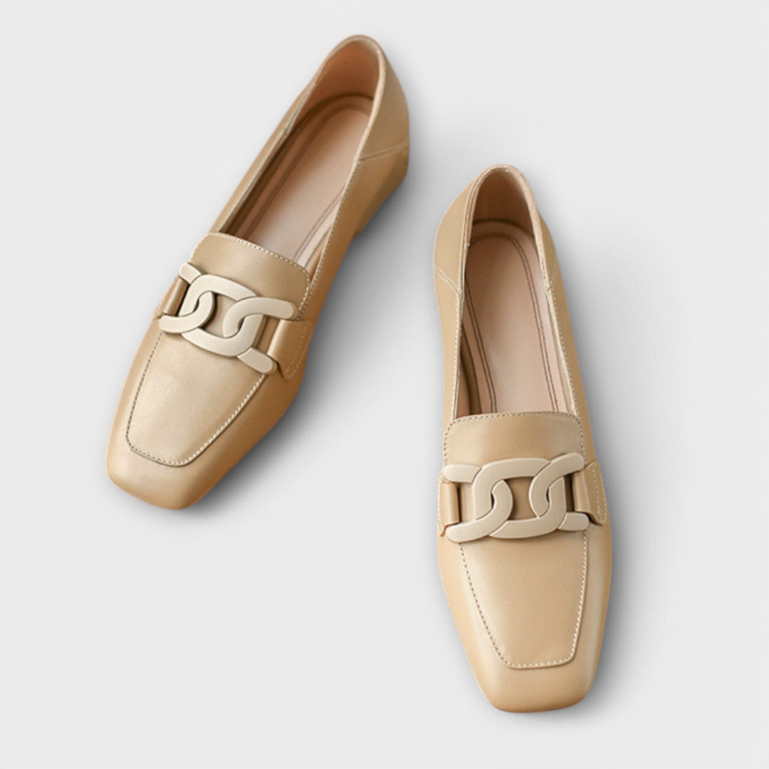 A pair of soft beige square-toe loafers with a chain-link ornament across the front.