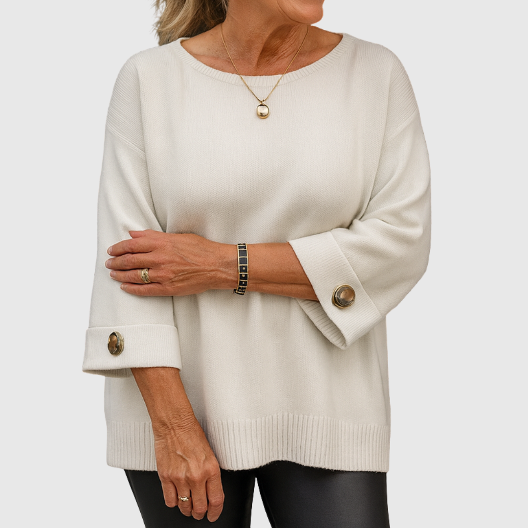 A woman wearing a cream sweater with a wide round neck and gold-toned buttons on the cuffs.