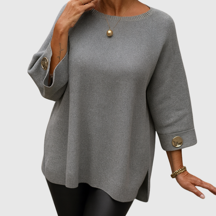 A woman wearing a cream sweater with a wide round neck and gold-toned buttons on the cuffs.