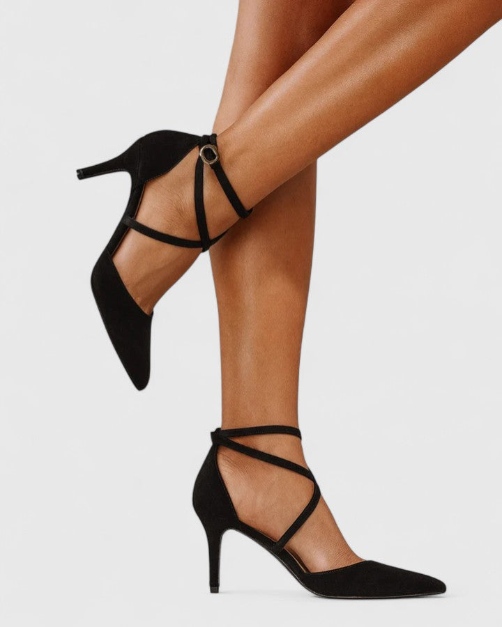 A woman wearing black pointed-toe heels with crossover ankle straps and a slim stiletto heel, dressed for an evening out.