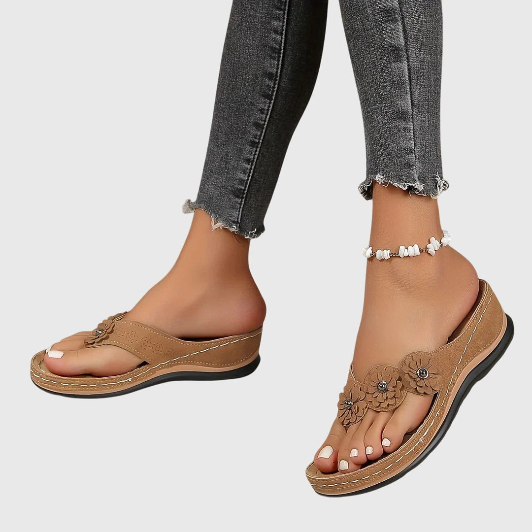 A woman wearing tan flower-trim flip-flops with a toe-post front and a low wedge sole.