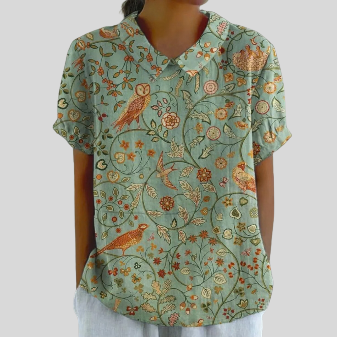 A woman wearing a flowing polo collar top in soft sage with a bird-and-botanical print of owls and a pheasant.