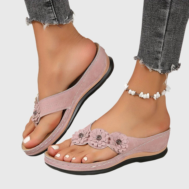 A woman wearing tan flower-trim flip-flops with a toe-post front and a low wedge sole.