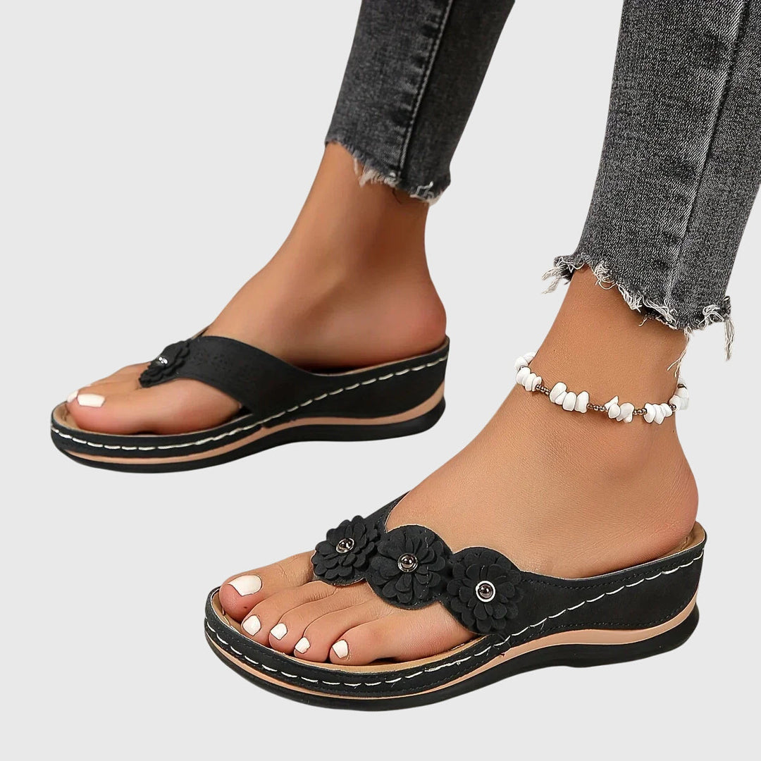 A woman wearing tan flower-trim flip-flops with a toe-post front and a low wedge sole.