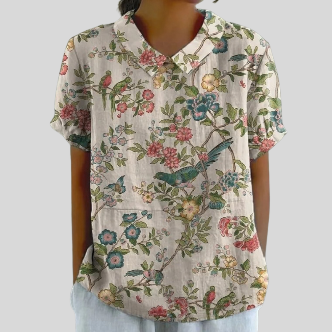 A woman wearing a cream top printed with songbirds and blossoming branches, finished with a refined collar, at a relaxed lunch with friends.