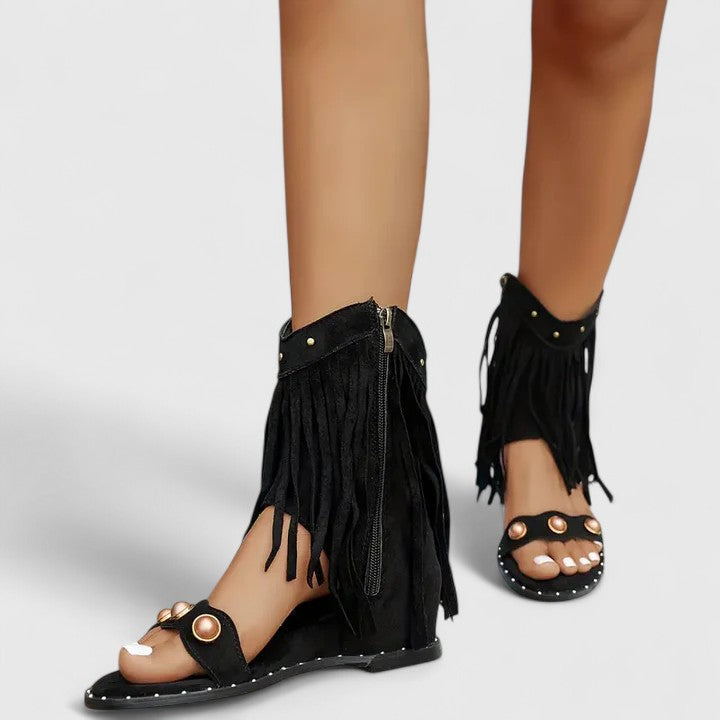 A woman wearing khaki fringe wedge sandals with a fringed ankle cuff and a back zip.