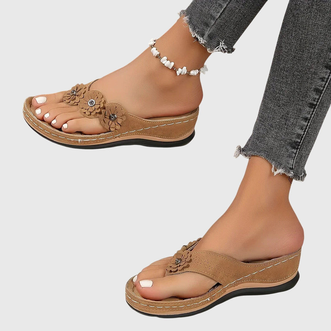 A woman wearing tan flower-trim flip-flops with a toe-post front and a low wedge sole.