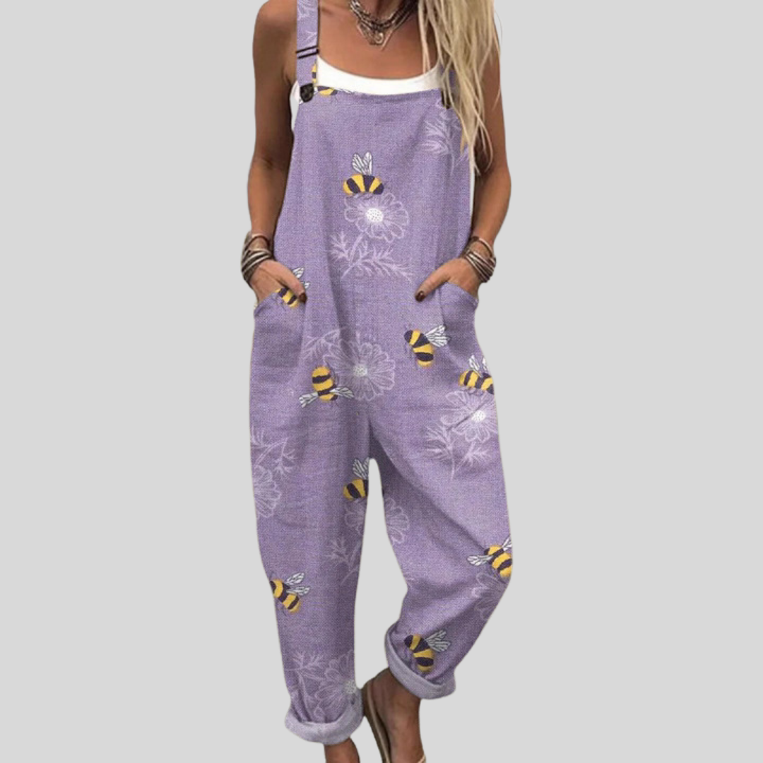 A woman wearing soft lilac overalls printed with golden bees and dandelion clocks, smiling on a bright day.