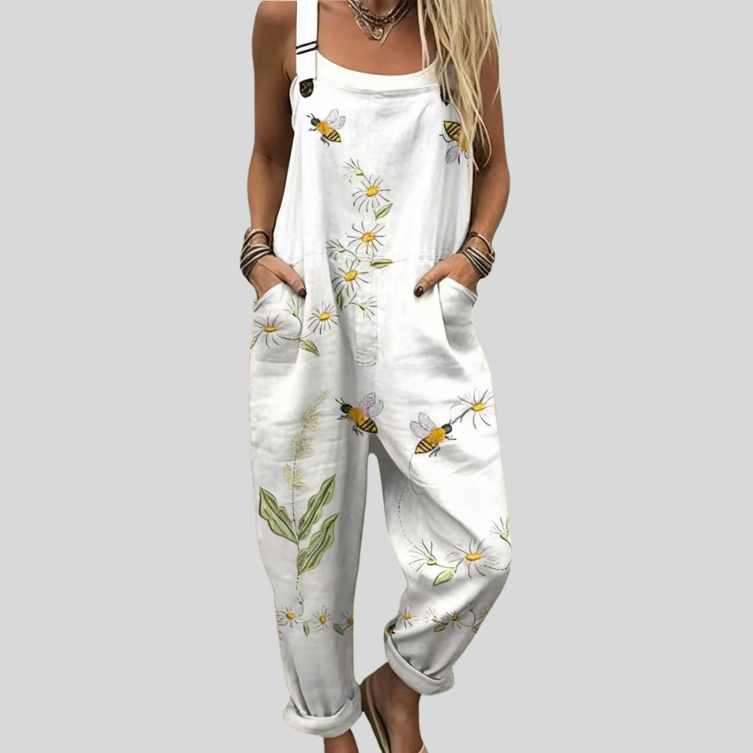 Woman wearing white overalls printed with daisies and little bees, standing relaxed in a summer garden.