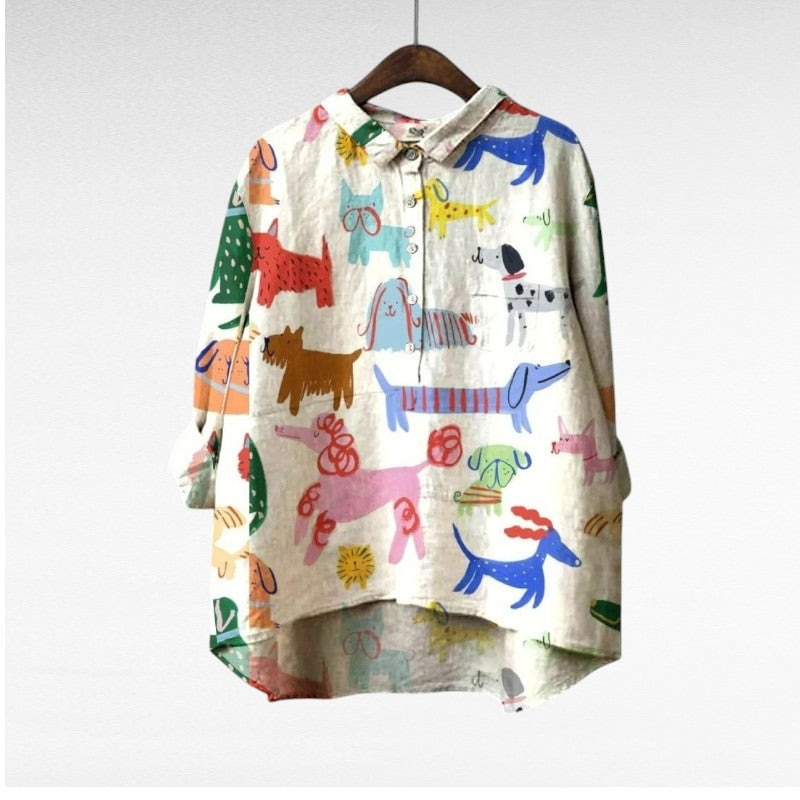 A relaxed blue button-up blouse printed with playful brown cartoon dachshunds and a small flower, shown on a hanger.