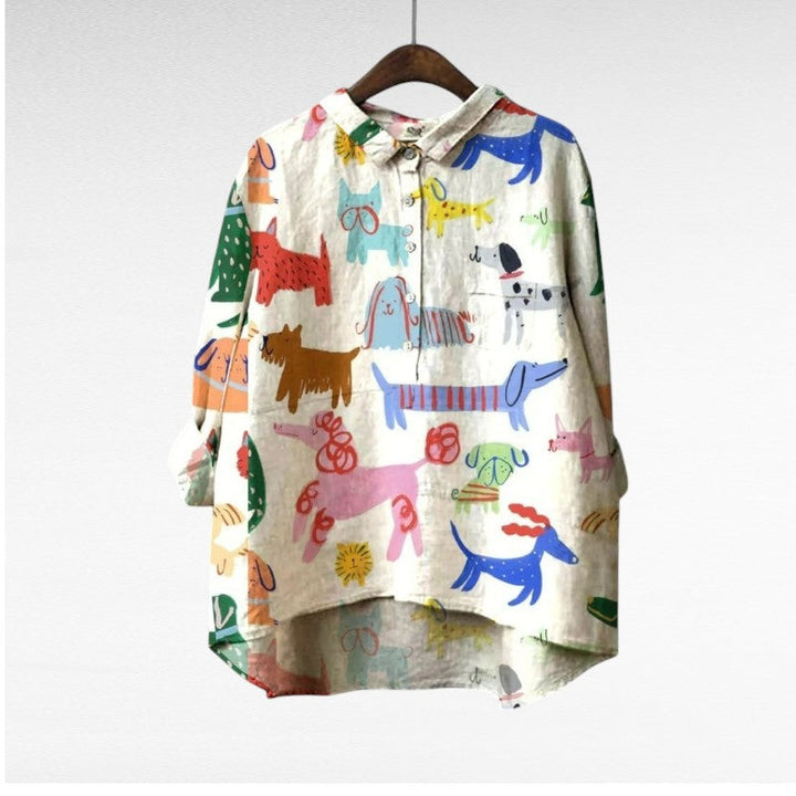A relaxed blue button-up blouse printed with playful brown cartoon dachshunds and a small flower, shown on a hanger.