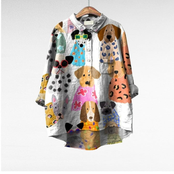 A relaxed blue button-up blouse printed with playful brown cartoon dachshunds and a small flower, shown on a hanger.