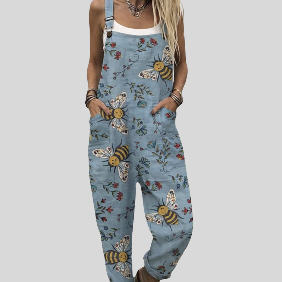 Woman wearing light blue overalls printed with bees, butterflies and fine flowers, standing relaxed and smiling.