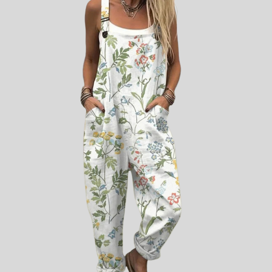 Woman wearing white overalls printed with delicate wildflowers and grasses in soft colours, standing relaxed.