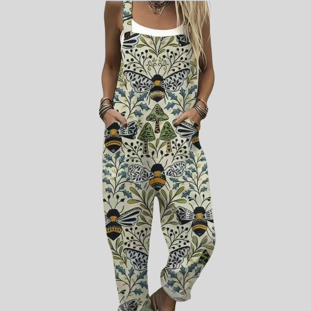 A woman wearing cream overalls with a symmetrical folk-art print of bees, mushrooms and green leaves, relaxed outdoors.