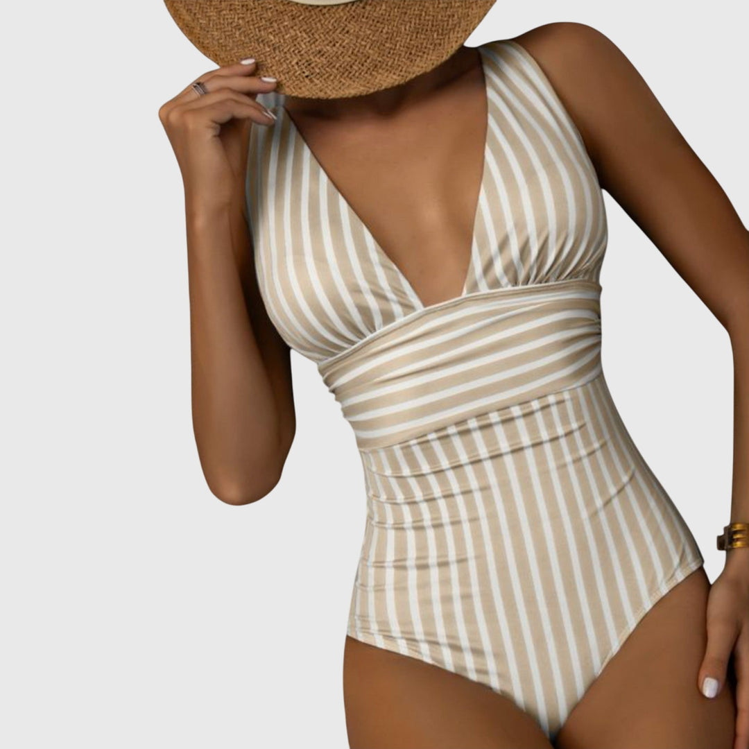 A woman by the sea in a beige and white striped V-neck swimsuit, relaxed and glowing in the sun.