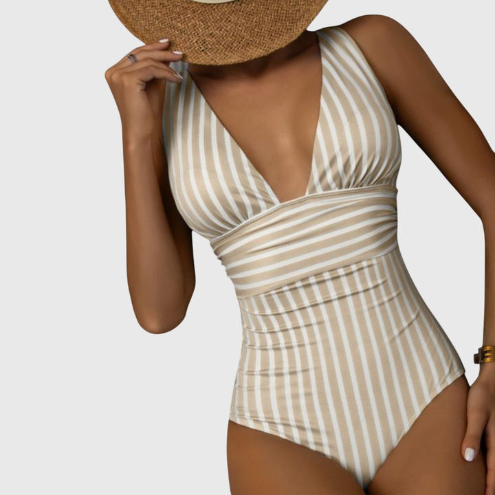 A woman by the sea in a beige and white striped V-neck swimsuit, relaxed and glowing in the sun.