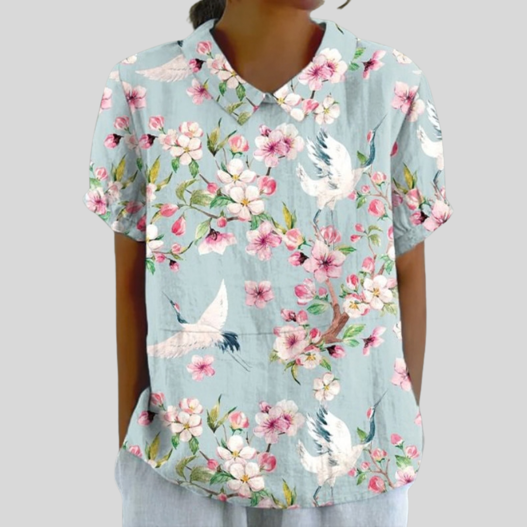 A woman wearing a flowing pale-blue polo collar top with a crane-and-blossom print, looking feminine and self-assured.