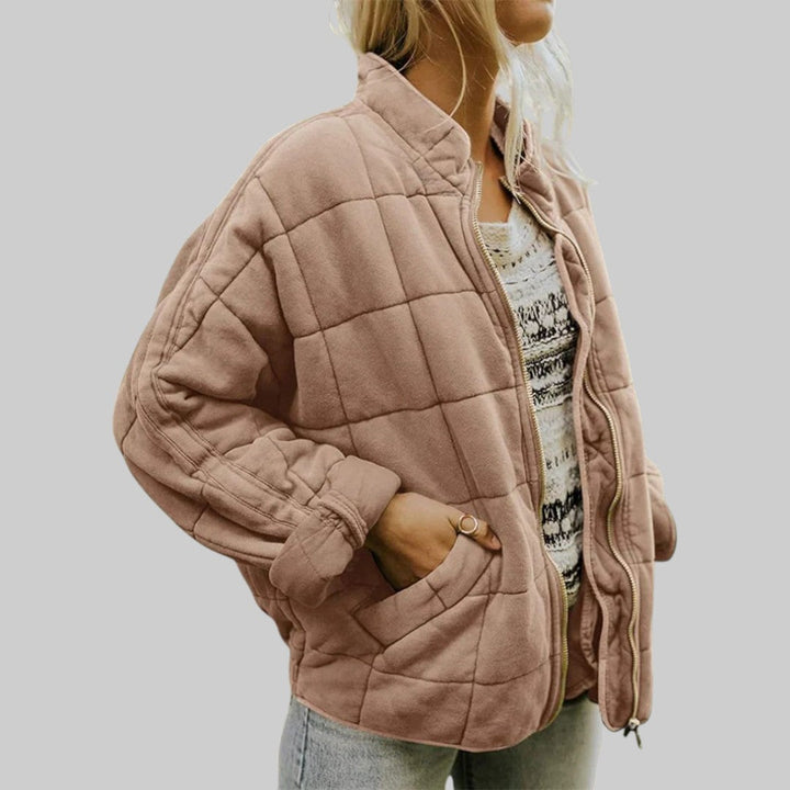 A woman wearing an oversized blush pink quilted jacket with a stand collar and zip front, hand in the pocket.