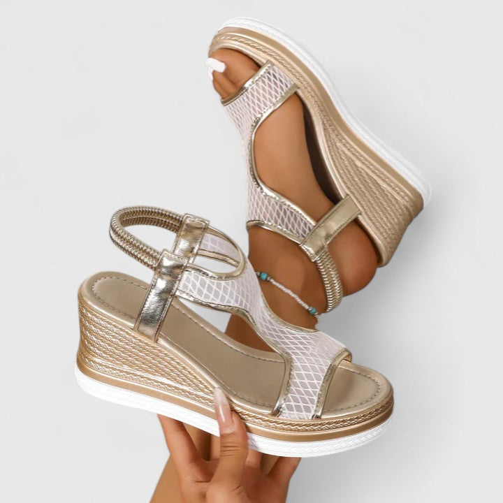 A woman wearing golden platform wedge sandals with a T-bar front, open-weave panels and a stretchy slingback.