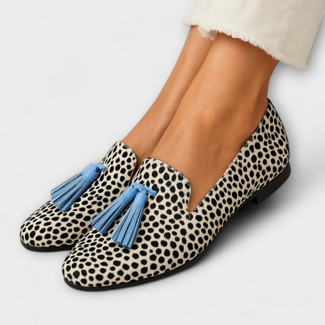 A woman wearing cream and black spotted print slip-on loafers with bright blue tassels, styled with jeans.