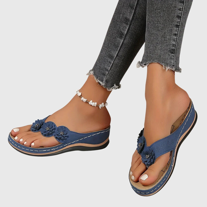 A woman wearing tan flower-trim flip-flops with a toe-post front and a low wedge sole.