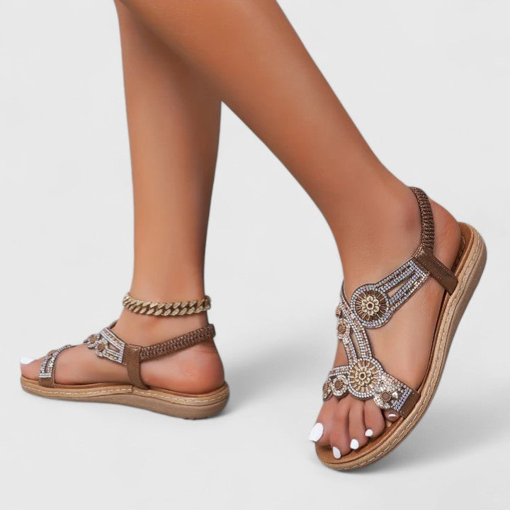 A woman wearing brown embellished sandals with sparkling stones, golden medallions and a stretchy slingback.
