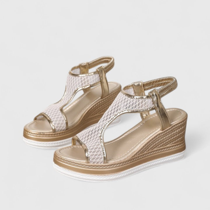 A woman wearing golden platform wedge sandals with a T-bar front, open-weave panels and a stretchy slingback.