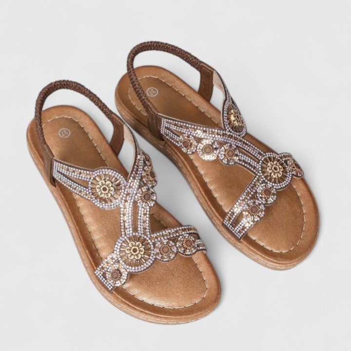 A woman wearing brown embellished sandals with sparkling stones, golden medallions and a stretchy slingback.