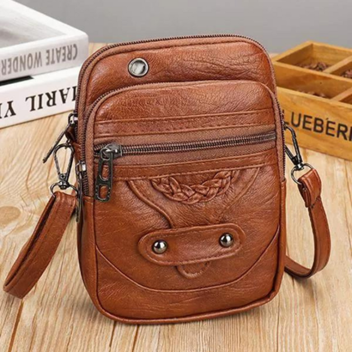 A small brown crossbody bag with a plaited front detail and zip pockets on a wooden stump.
