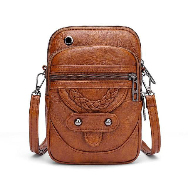 A small brown crossbody bag with a plaited front detail and zip pockets on a wooden stump.