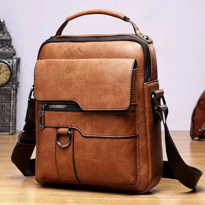 A tan crossbody bag with a flap front, zip pocket and wide adjustable strap on a wooden stump.