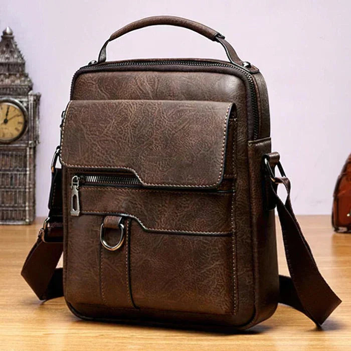 A tan crossbody bag with a flap front, zip pocket and wide adjustable strap on a wooden stump.