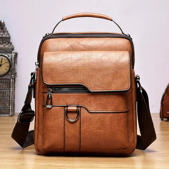 A tan crossbody bag with a flap front, zip pocket and wide adjustable strap on a wooden stump.