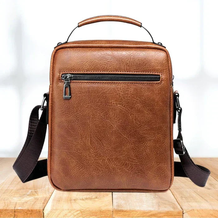 A tan crossbody bag with a flap front, zip pocket and wide adjustable strap on a wooden stump.