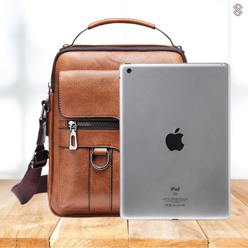 A tan crossbody bag with a flap front, zip pocket and wide adjustable strap on a wooden stump.
