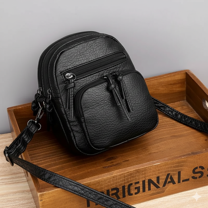 A small black everyday crossbody bag with zip pockets and an adjustable strap on a wooden stump.