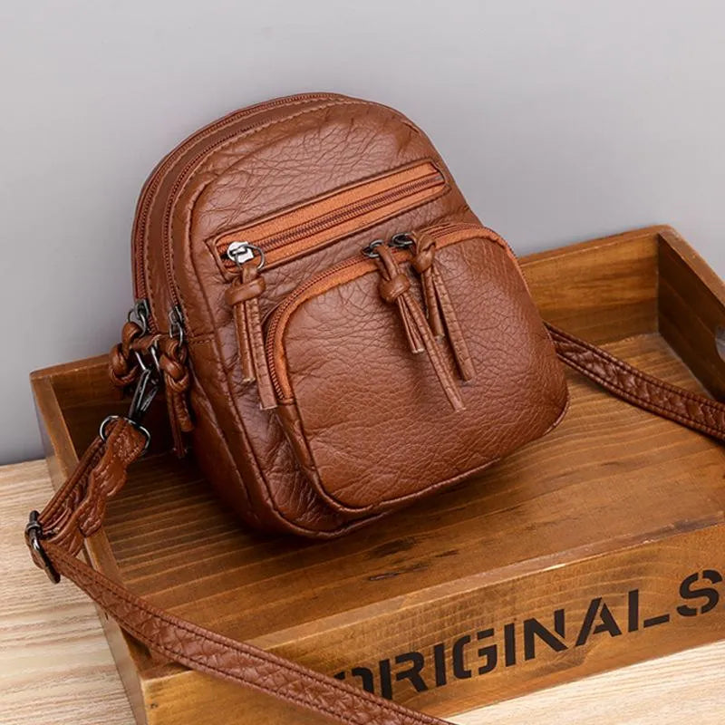 A small black everyday crossbody bag with zip pockets and an adjustable strap on a wooden stump.