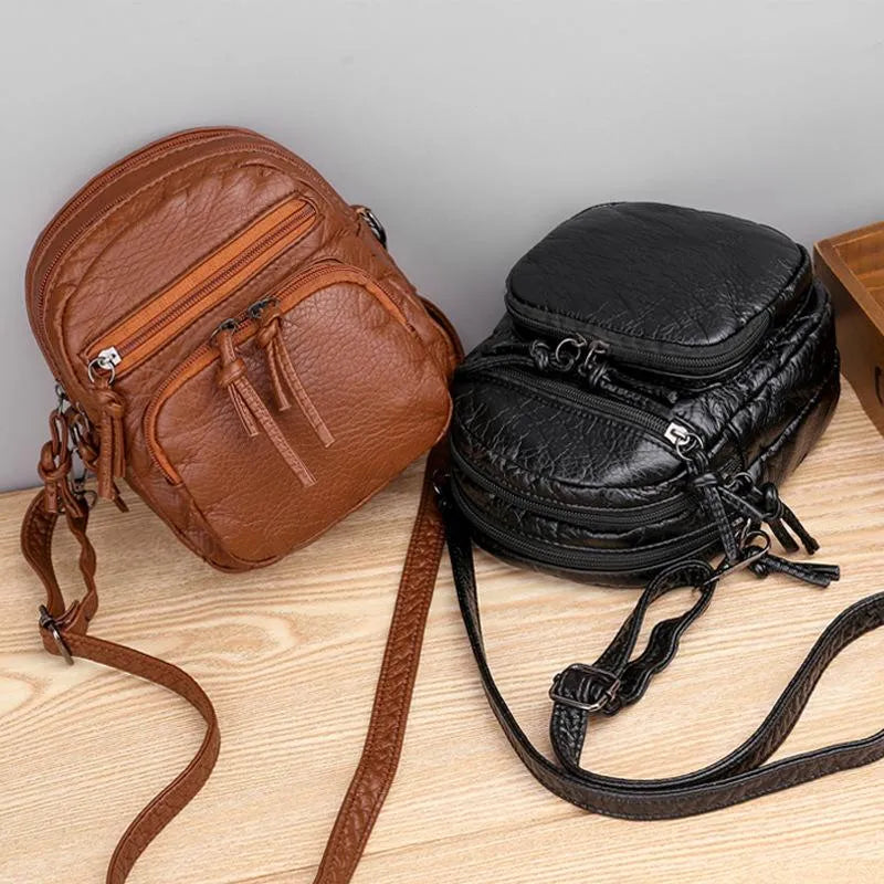 A small black everyday crossbody bag with zip pockets and an adjustable strap on a wooden stump.