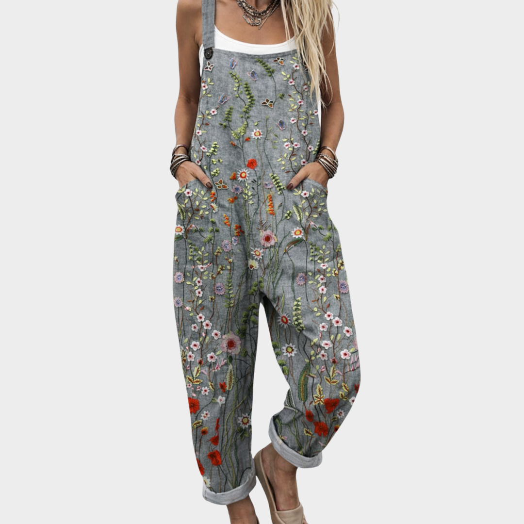 A woman wearing grey overalls covered in bright meadow flowers, daisies and poppies, with adjustable straps.