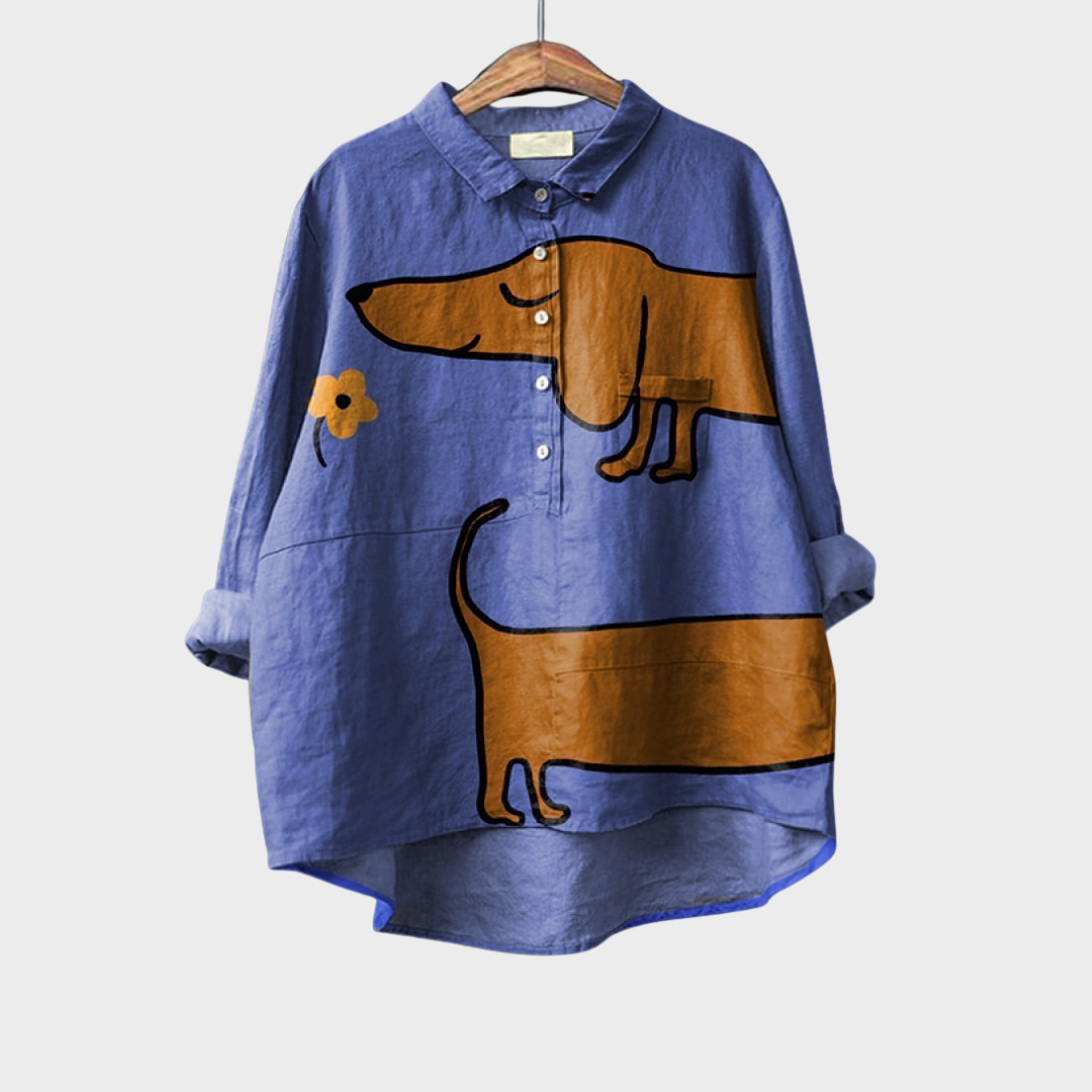 A relaxed blue button-up blouse printed with playful brown cartoon dachshunds and a small flower, shown on a hanger.