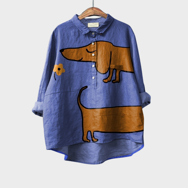 A relaxed blue button-up blouse printed with playful brown cartoon dachshunds and a small flower, shown on a hanger.