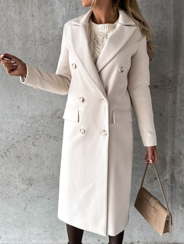 A woman wearing a long waisted coat, standing elegantly outdoors