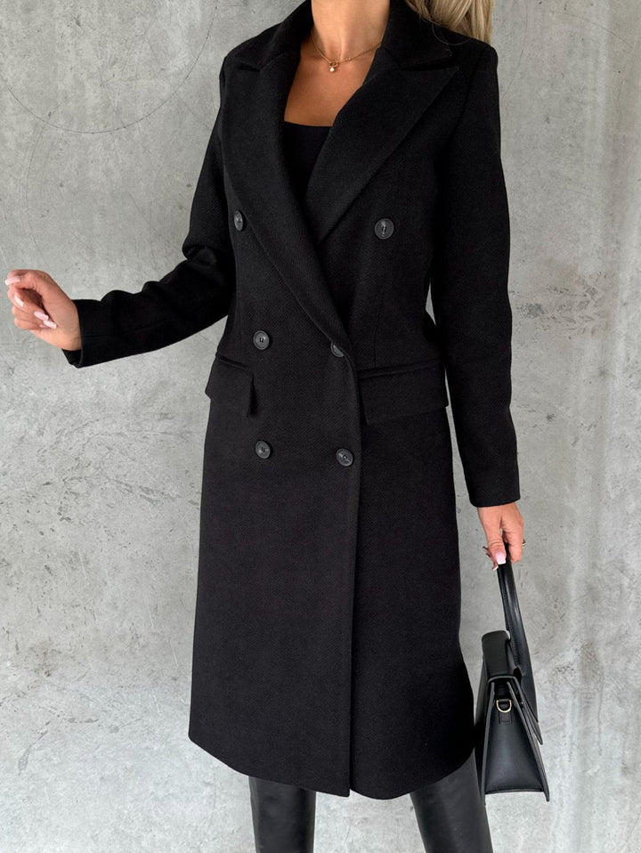 A woman wearing a long waisted coat, standing elegantly outdoors
